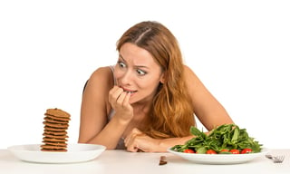 Portrait young woman deciding whether to eat healthy food or sweet cookies she is craving sitting at table isolated white background. Human face expression emotion reaction. Diet nutrition concept.jpeg
